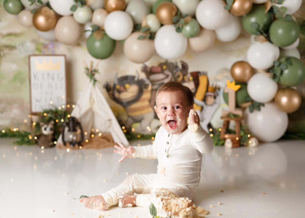 "Where the Wild Things Are" themed cake smash photoshoot in West Palm Beach, FL. Baby boy in white onesie has cake on his hands and feet and yelling at the camera. There is a "Where the Wild Things Are" backdrop, green, gold, and cream balloon arch, and canvas tent.