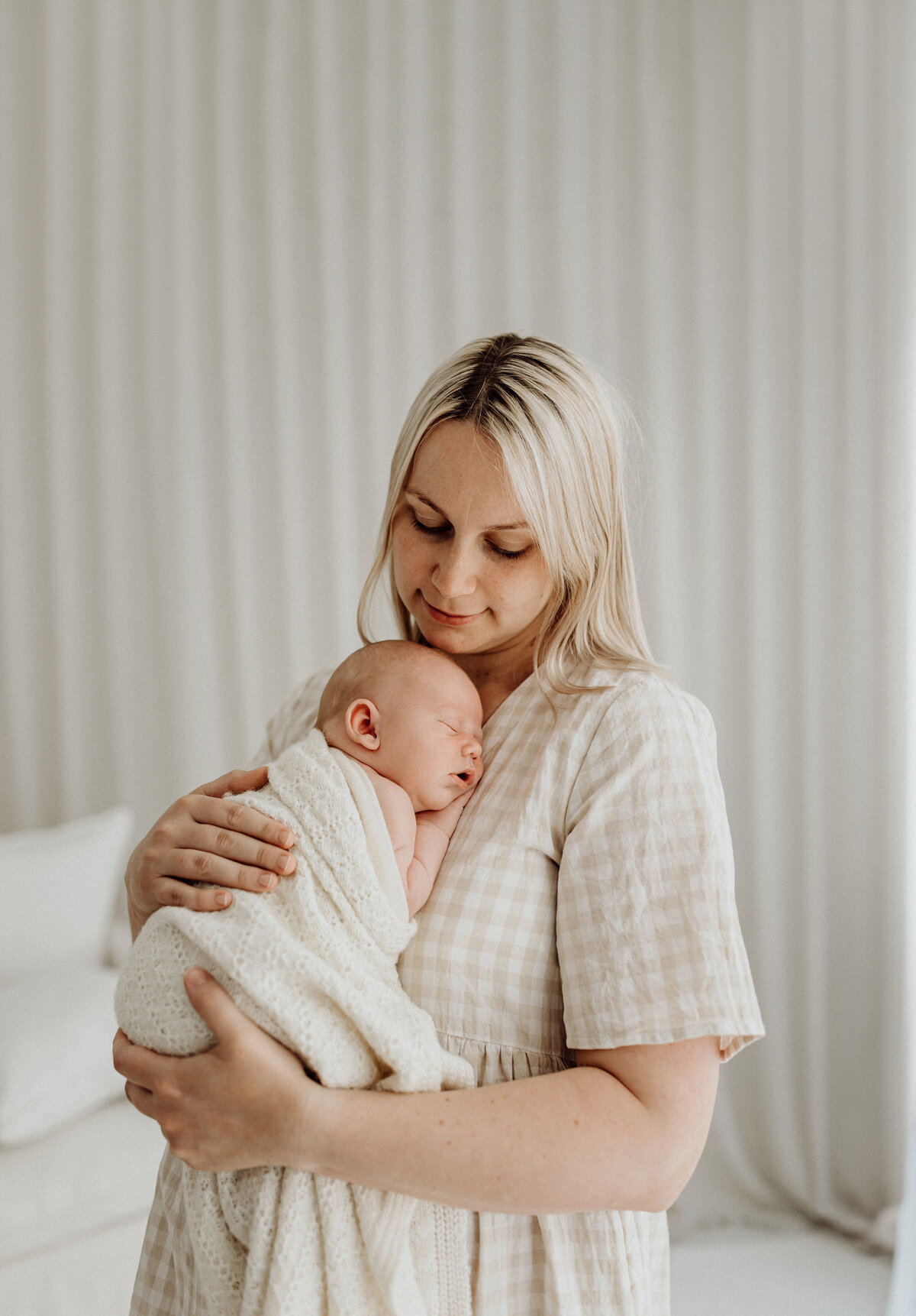 A mother holds her newborn baby wearing a white and beige dress with a sheer curtain as the backdrop in lucy janes photography studio in christchurch