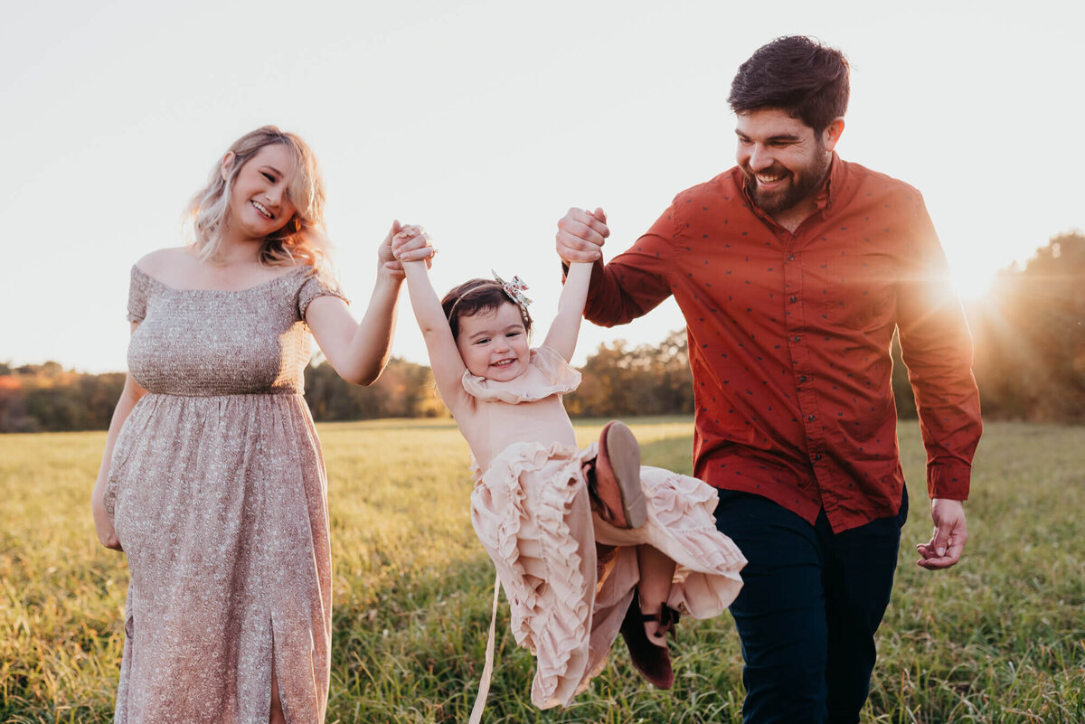A mother and father swing their toddler daughter between them at sunset in a field