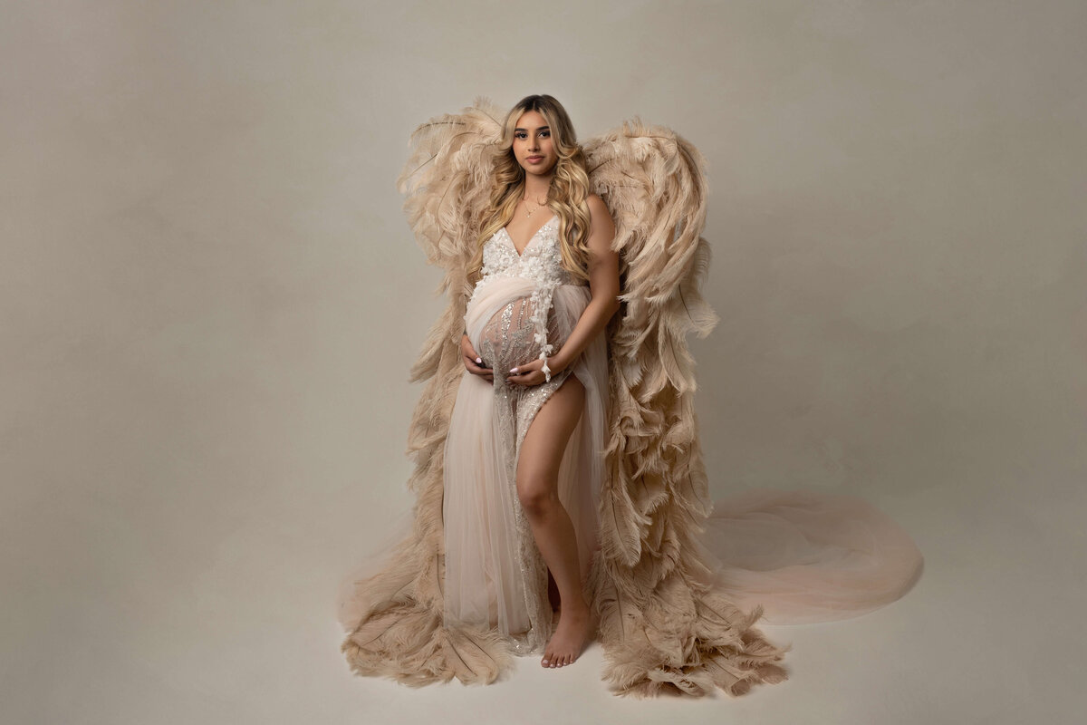 A mother to be stands in a studio wearing a white maternity gown and holds her bump with large feather wings