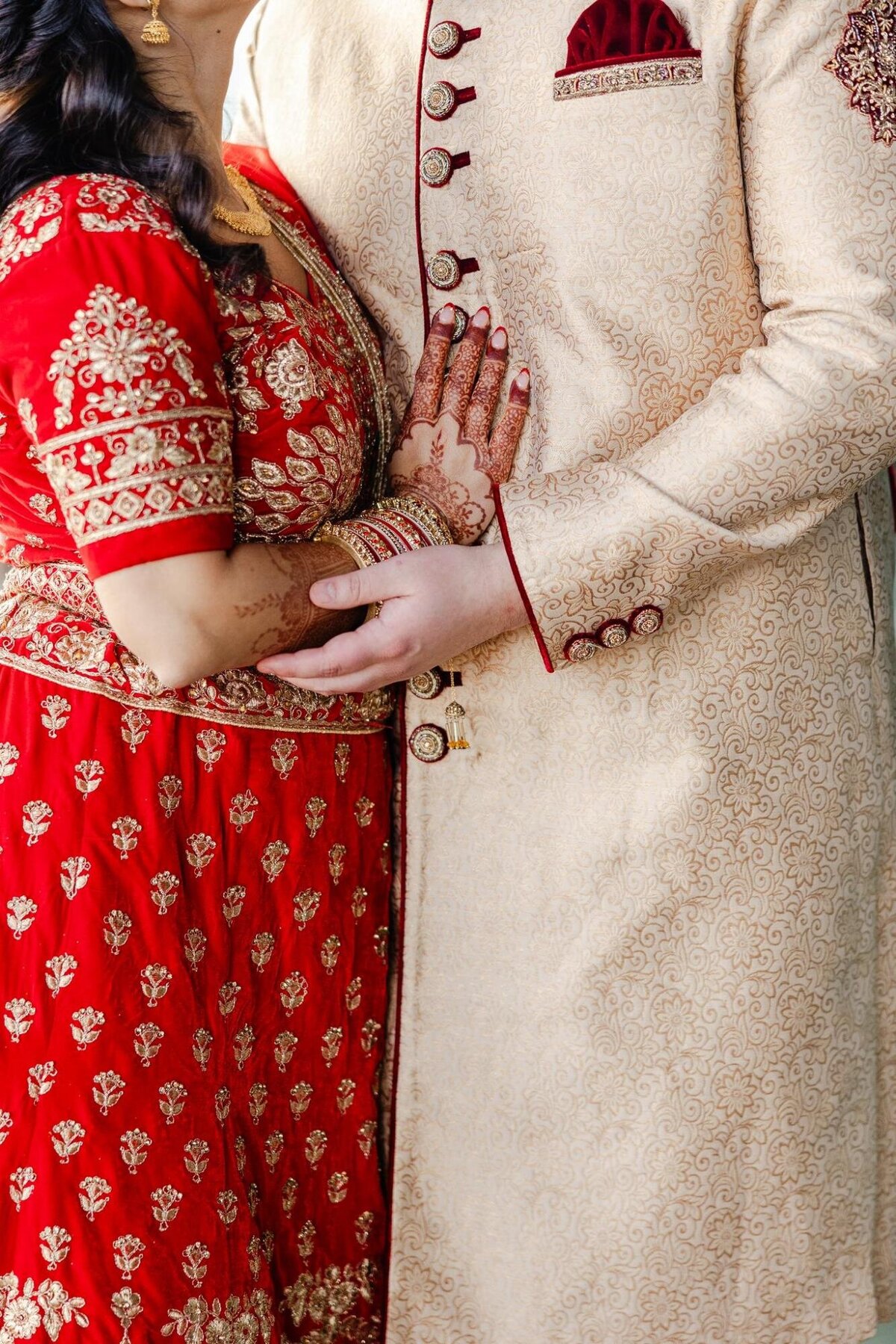 A bride in a red embroidered dress and henna on her hands holds onto a groom wearing a cream-colored, ornate sherwani.