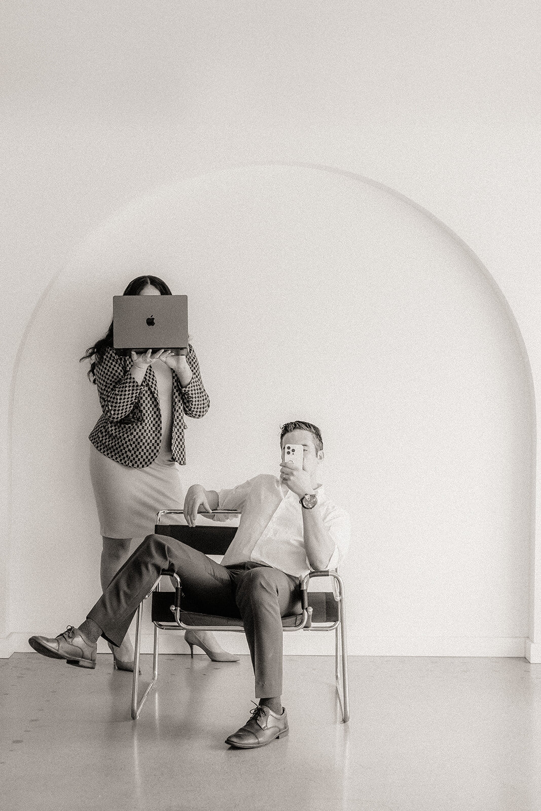 A man holds a phone in a chair while a woman stands behind on a laptop