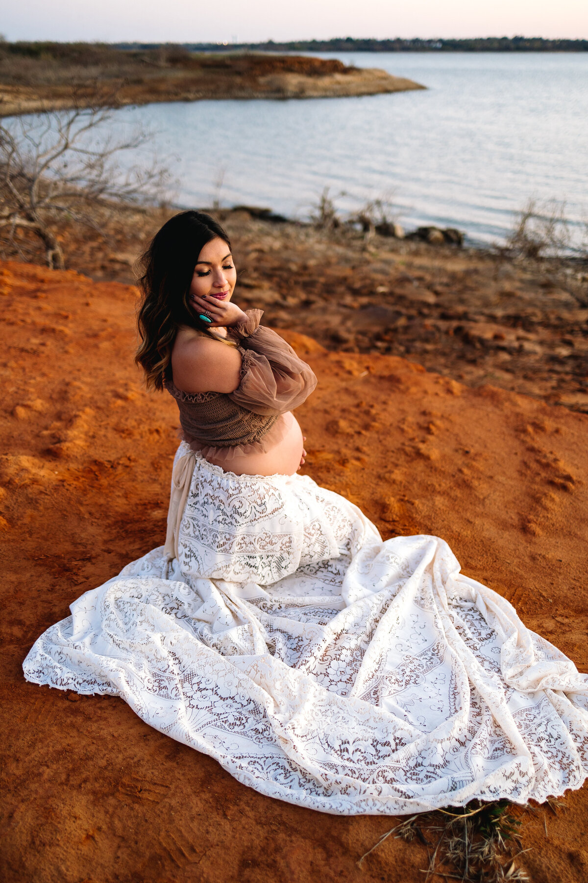 Maternity moments affordably with professional photo sessions in Albuquerque. The image features a pregnant woman elegantly dressed in a white lace skirt and brown blouse, sitting peacefully on the sand by a lake. Her hand rests on her cheek, and her eyes are closed, showcasing a serene and beautiful maternity pose.