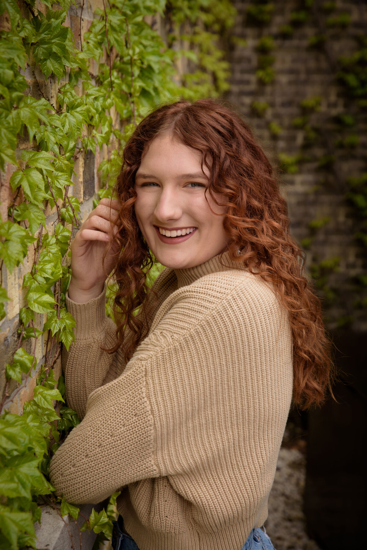 Green Bay East High School senior girl sitting wearing a tan sweater and jeans leaning against a tan brick wall with green leafy vines in urban setting in downtown Green Bay, Wisconsin.