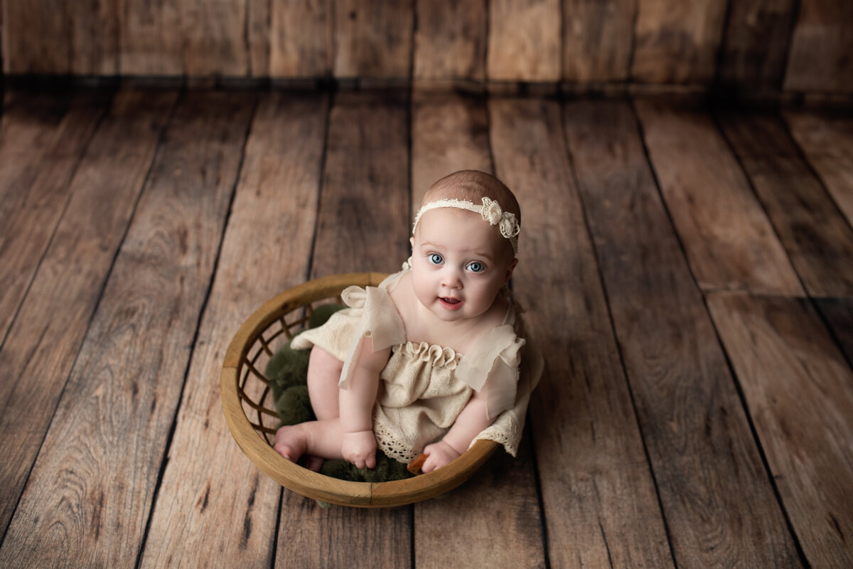Milestone baby girl portrait in vintage basket with wooden backdrop—styled with heirloom textures and charm.