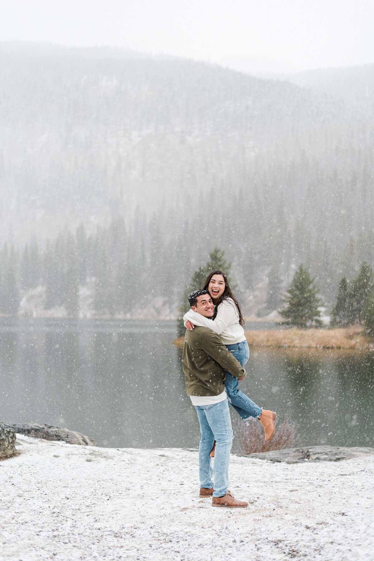 denver couples photography during winter with man picking up woman while it snows on a lake behind them in the woods captured by denver engagement photographer
