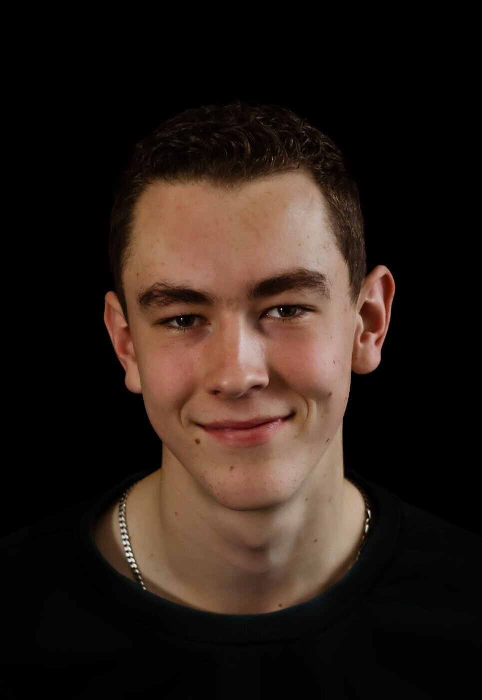 Een portretfoto van een jonge man met een open en vriendelijke uitstraling, gefotografeerd tegen een donkere achtergrond