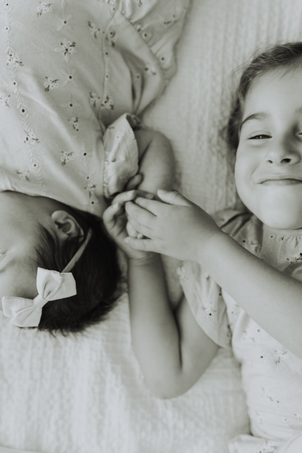Black and white photo of a little girl laying beside her baby sister on the bed holding her hand taken by a langley family photographer