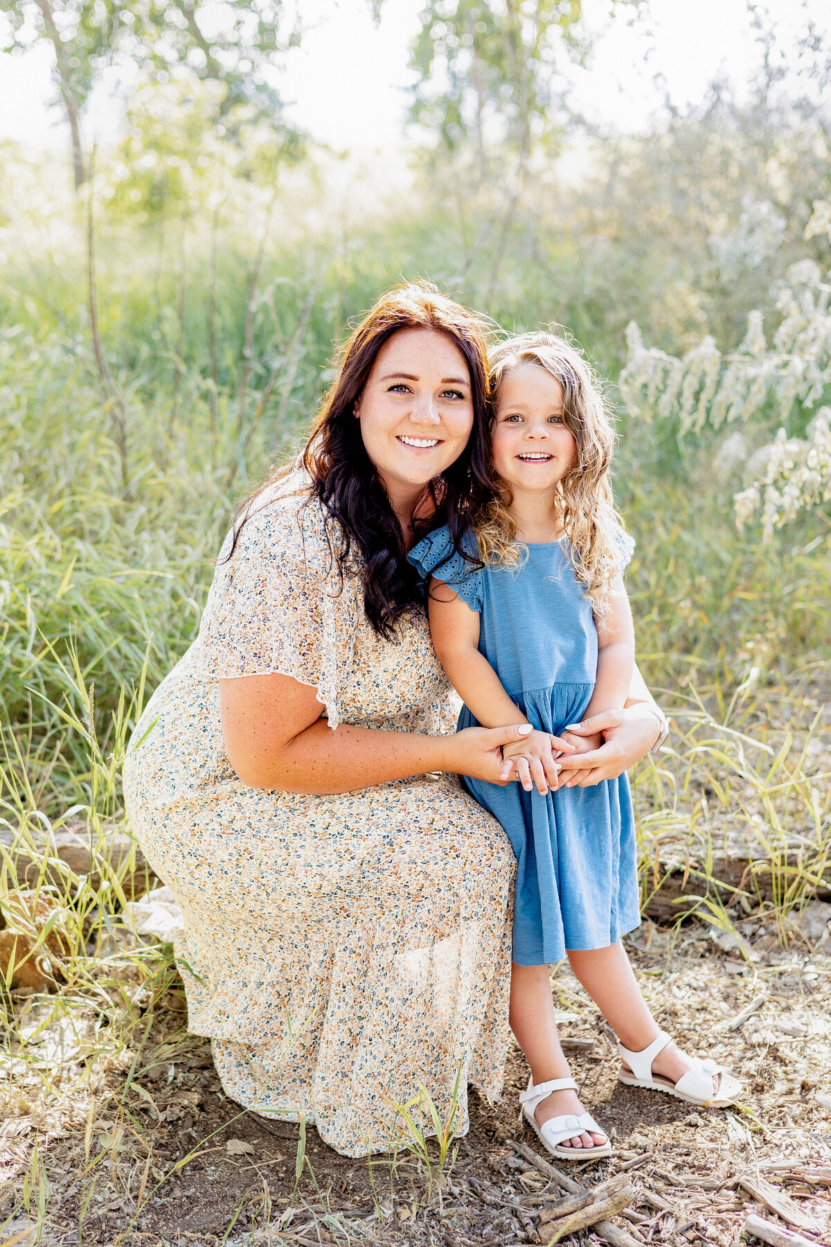 A mother with long dark hair in a floral dress kneels beside her young daughter in a blue dress as they hold hands and smile together in a grassy field.