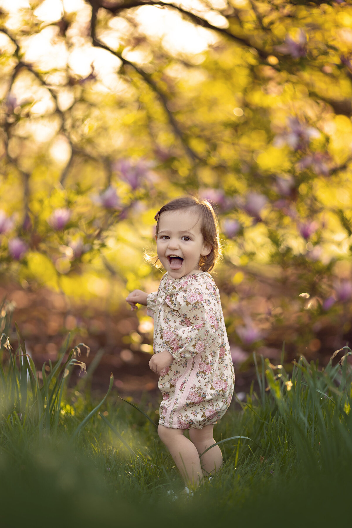 A toddler girl smiles while exploring a grassy lawn at sunset in a floral print dress