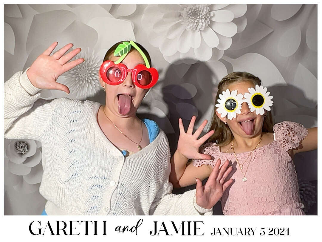two children wearing cherry and flowers sunglasses in front of a white backdrop in a photo booth capture