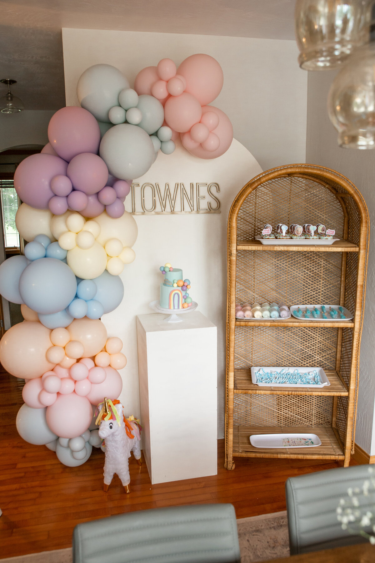 Pastel balloons set up on a white wooden backdrop, a rattan shelf with desserts displayed, and a white wooden cake stand in front.