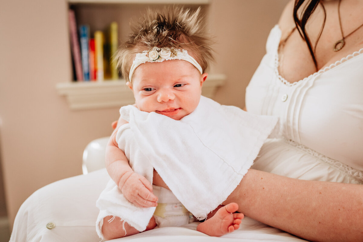 A newborn baby with spiky hair and a floral headband sits on a parent's lap.