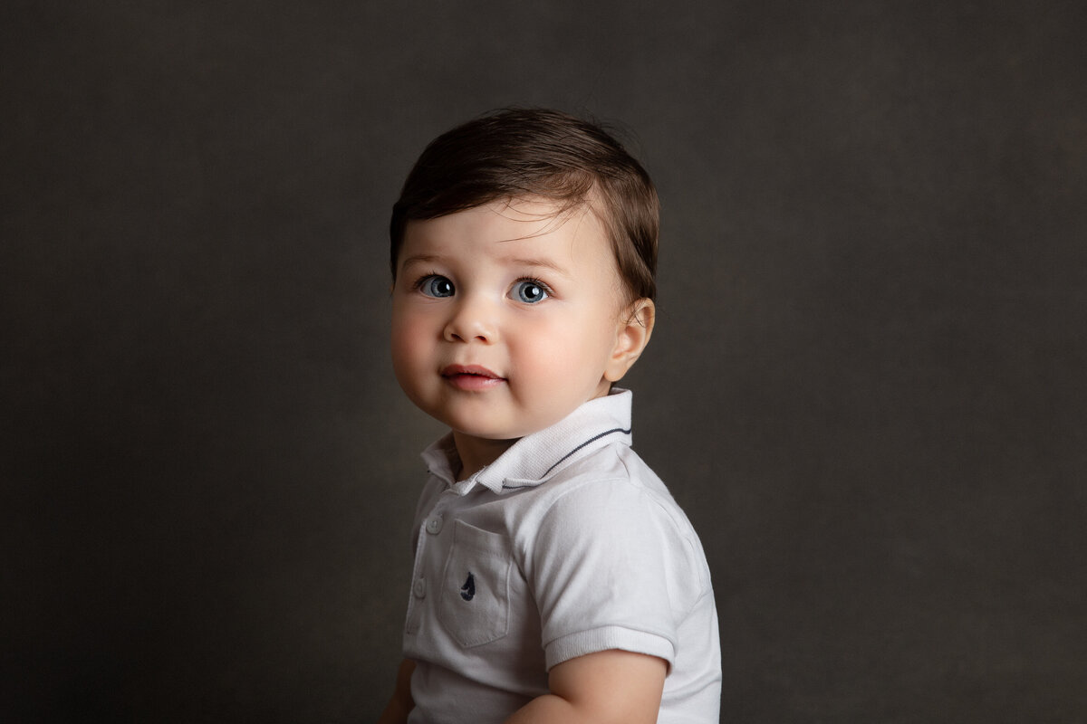 Six month old baby boy sitting sideways wearing a white polo shirt and looking at the camera smiling.