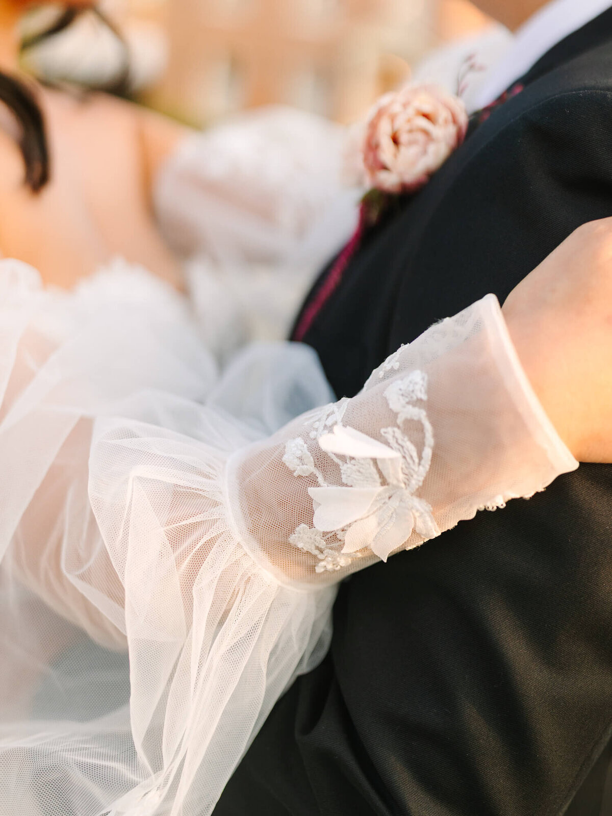 Close-up of a bride's lace sleeve and tulle fabric resting on a suited arm.
