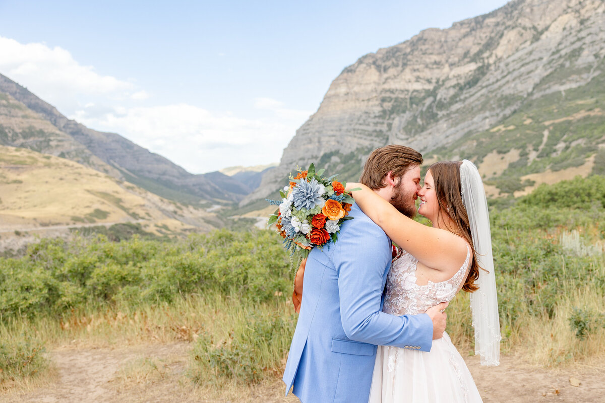 The bride and groom lean in for a romantic kiss with Provo Canyon’s towering mountains and lush greenery behind them. The bride’s bouquet of blue, orange, and white flowers adds a pop of color.