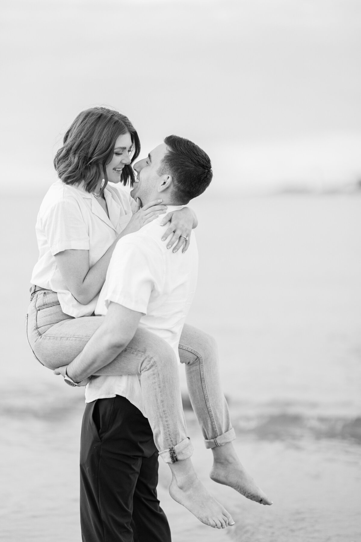 A black and white image of a man picking up and laughing with a woman during their Wingaersheek Beach sunset engagement photography session.