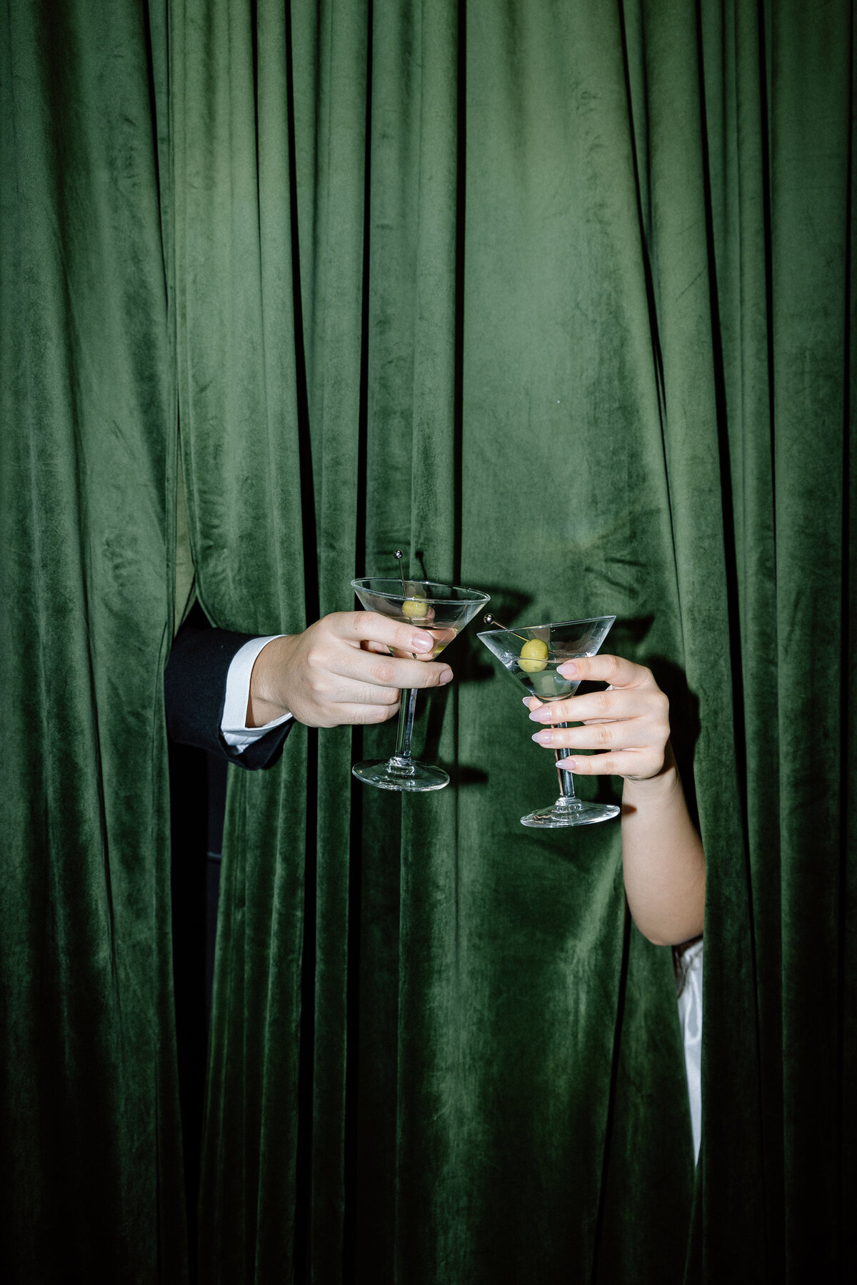 Two hands sticking out of a green velvet curtain toasting two martini glasses.
