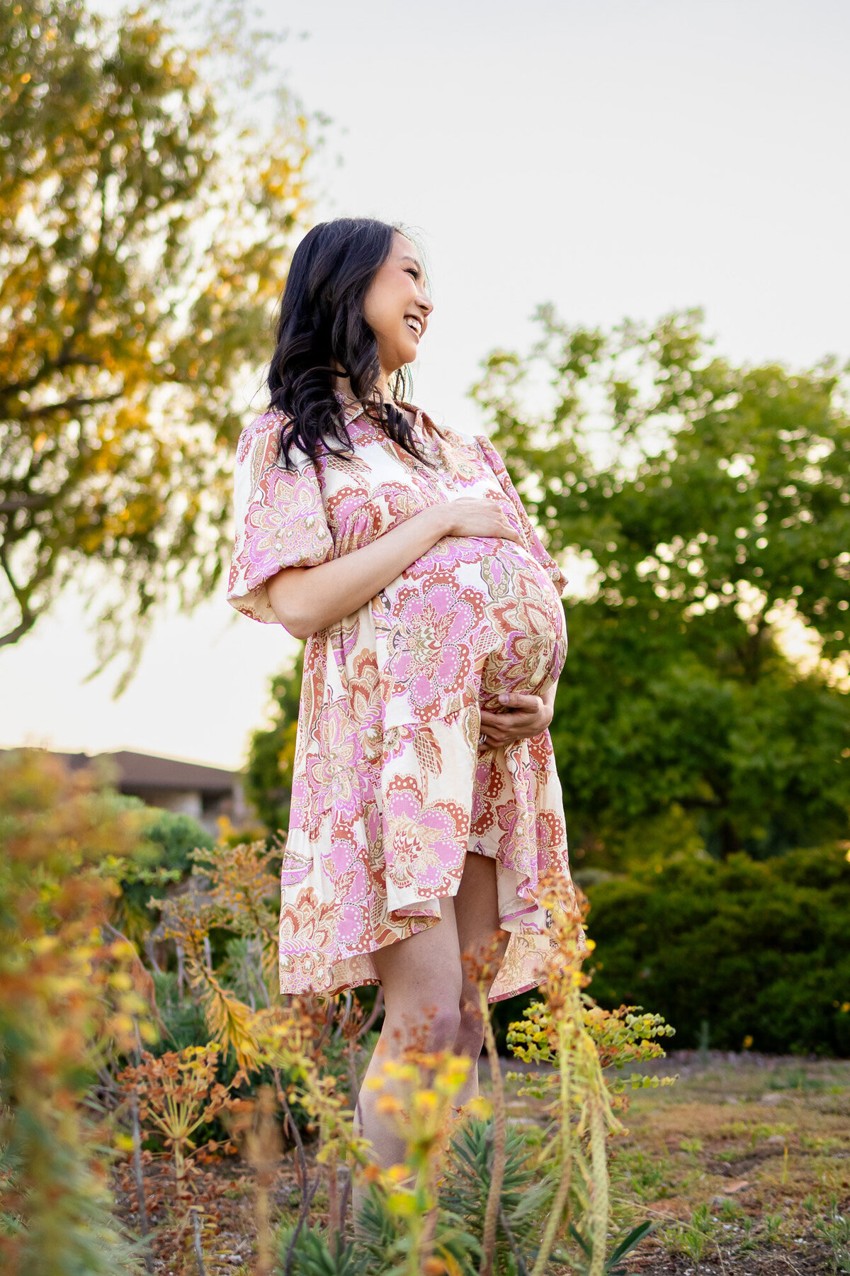 Radiant expecting mother smiles while holding her belly among soft green and yellow foliage — Bay Area Maternity Portfolio - Ellobelle Photography.