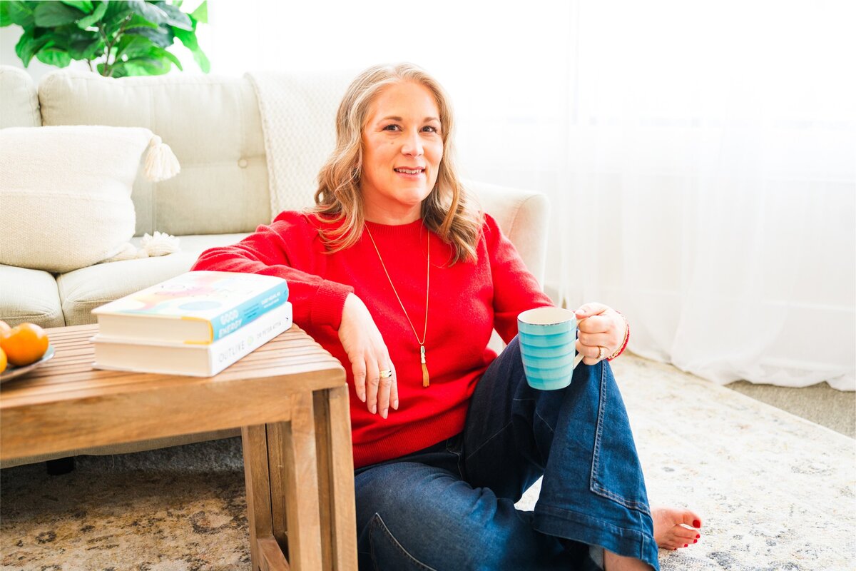 A middle aged female sitting on the floor leaning again a coffee table wearing a red sweater and holding a teal coffee mug, representing a relaxed vibe.