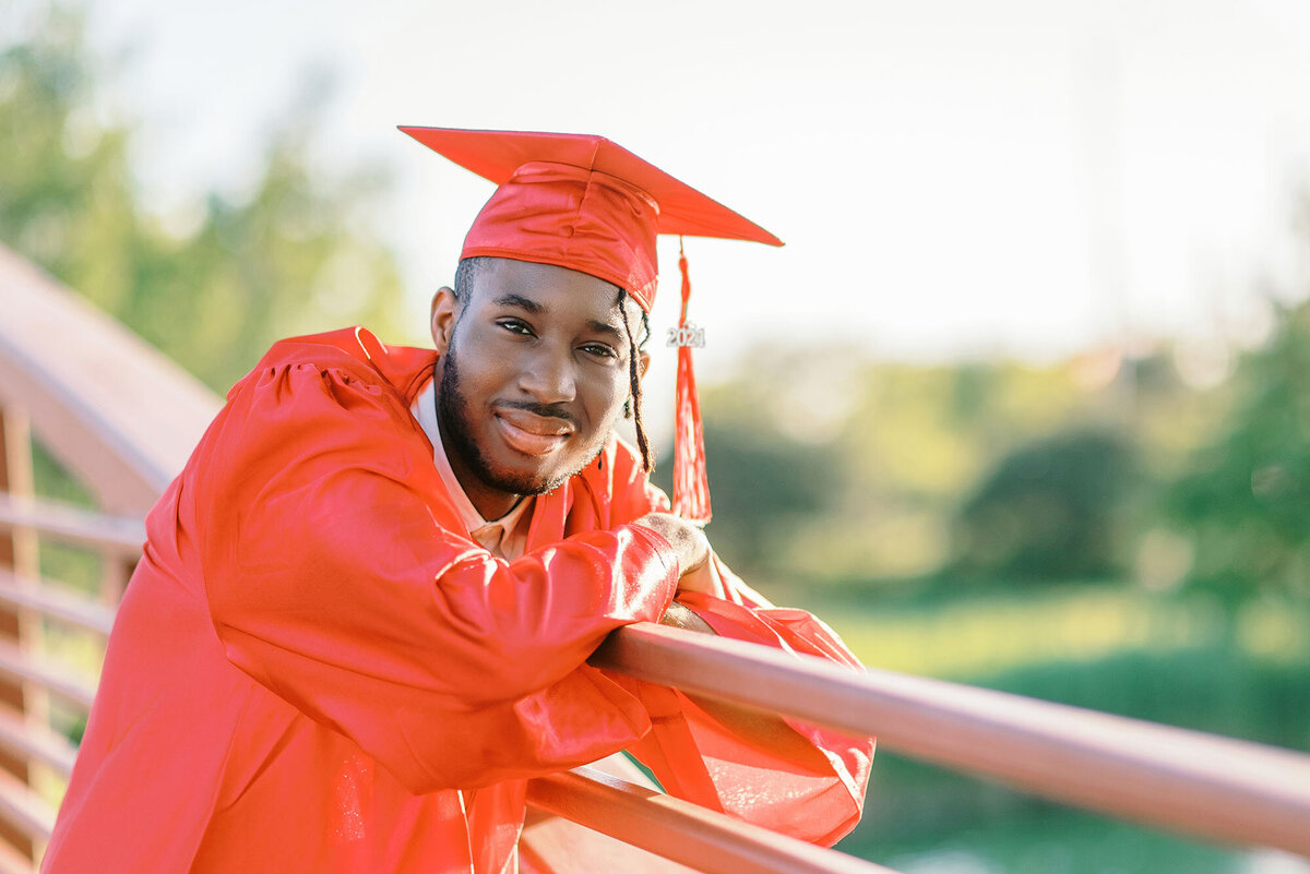 Cap and gown photos in Munster, IN - a male graduate in a red ca[ and gown leaning on a bridge in Centennial Park, Munster, IN