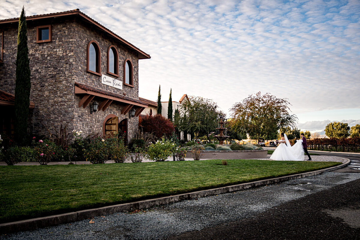 The groom lovingly adjusts the bride's train as they walk hand-in-hand into Casa Real, showcasing a sweet moment of care and partnership on their wedding day.