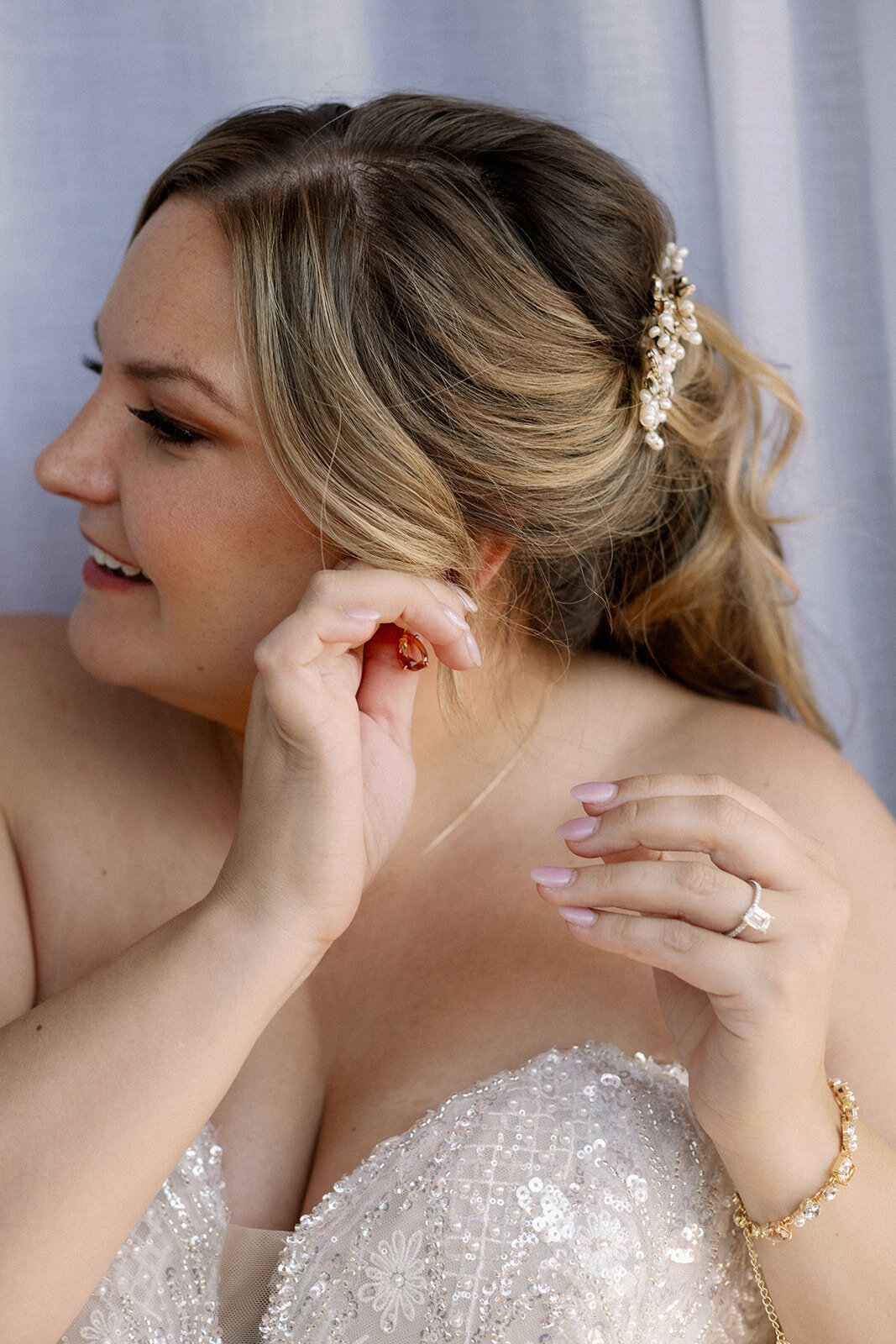 Bride puts on her earings on her wedding day in a strapless dress with a half up half down hairstyle with a gold clip.