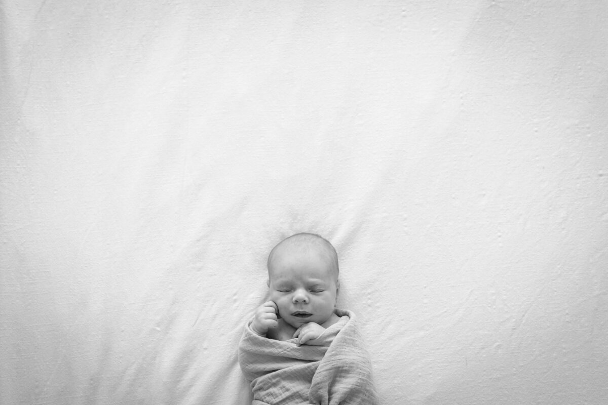 A black-and-white image of a newborn baby swaddled tightly in a blanket, peacefully sleeping on a bed with a soft, textured surface.
