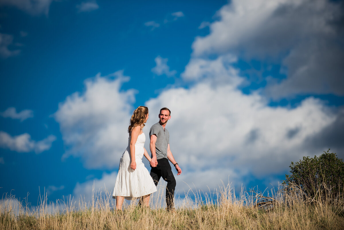 A wide angle shot of a man leading his fiancée through a golden field with a blue sky and clouds in the background.