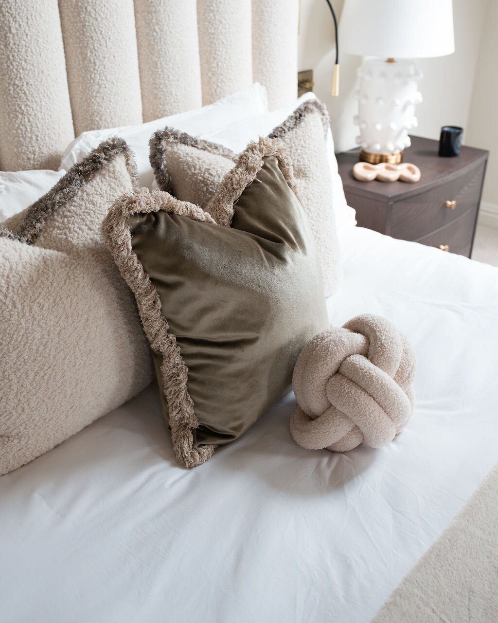 A bed with a white headboard, decorated with pristine white pillows, chic neutral throw pillows, and a plush beige knot pillow. A bedside table with a white lamp is visible in the background.