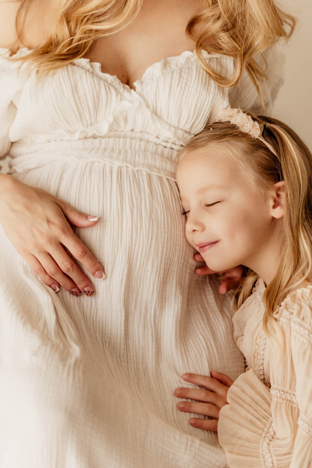 Older sister snuggles her mother's baby bump while closing her eyes and smiling.