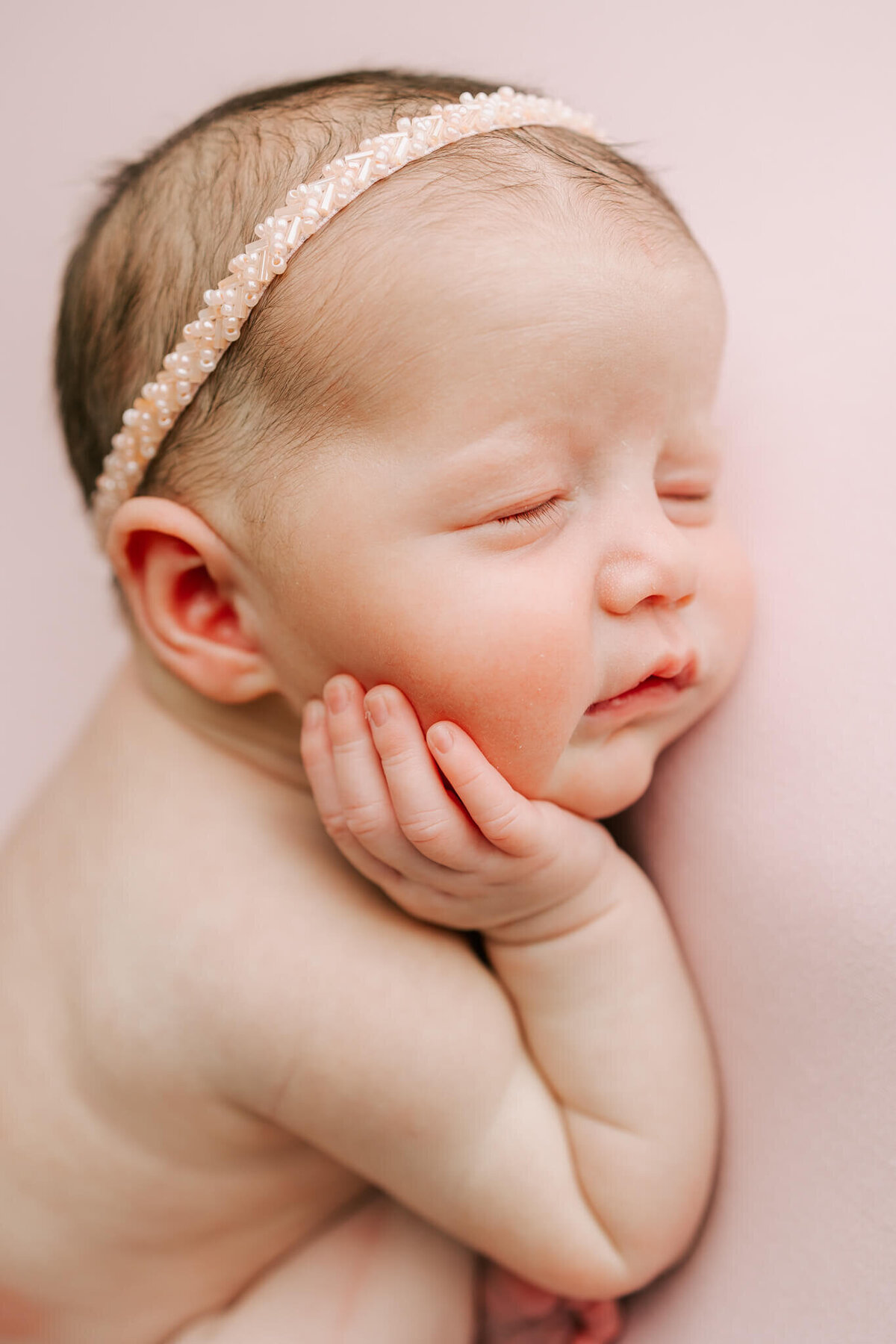 stunning portrait of a newborn baby girl with her hand on her cheek. She is wearing a pink headband