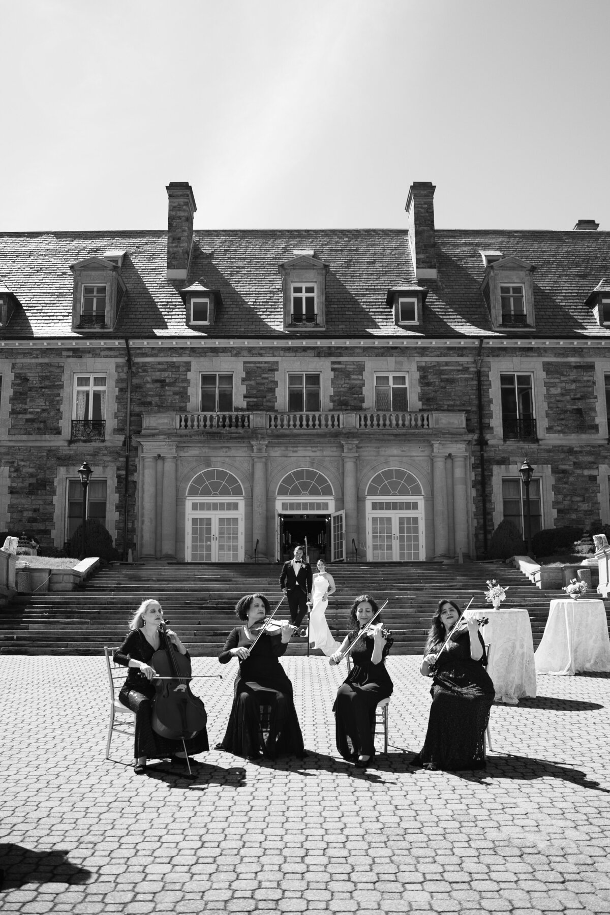 Rhode Island Wedding Photographers | Kelsey Sheehan Photography Timeless Rhode Island Weddings | Black and white image of a string quartet performing outdoors in front of a grand stone mansion. A couple stands on the stairs, creating an elegant ambiance.