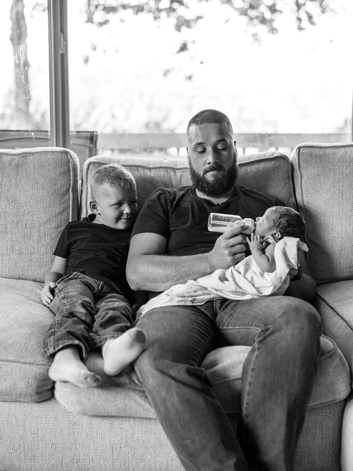 A man feeds a baby with a young boy sitting next to them during a newborn photography shoot held at their home.