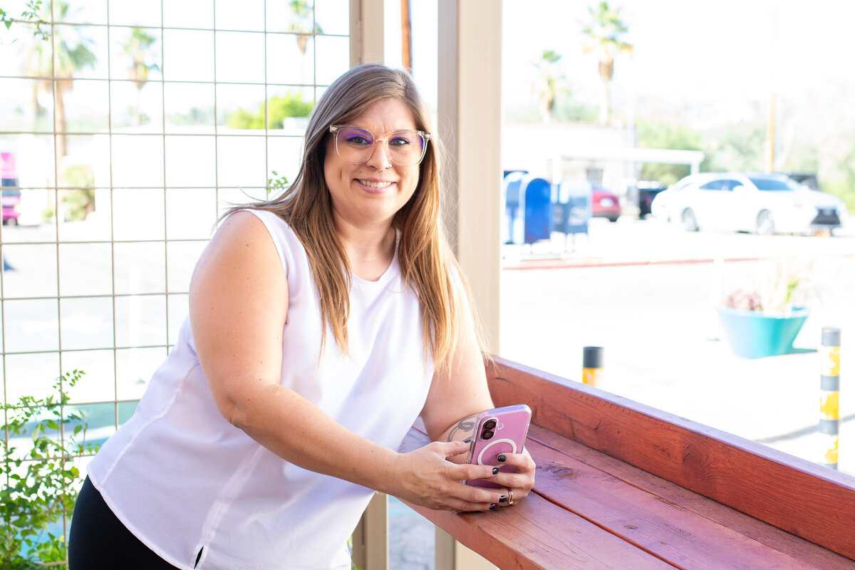 Smiling woman in a white sleeveless top holding a phone while leaning on a wooden counter outdoors, photographed by Vyrl Photo for Tucson brand photography.