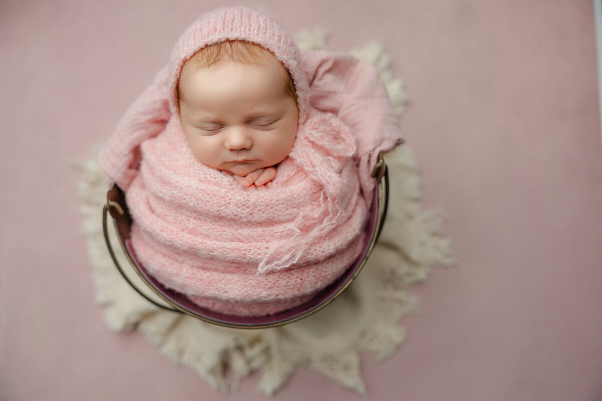 Blush-toned newborn portrait featuring a baby girl wrapped in textured pink knits, resting in a bucket against a soft pink background. Photographed by Inland Empire newborn photographer Kaitlyn Dawn.