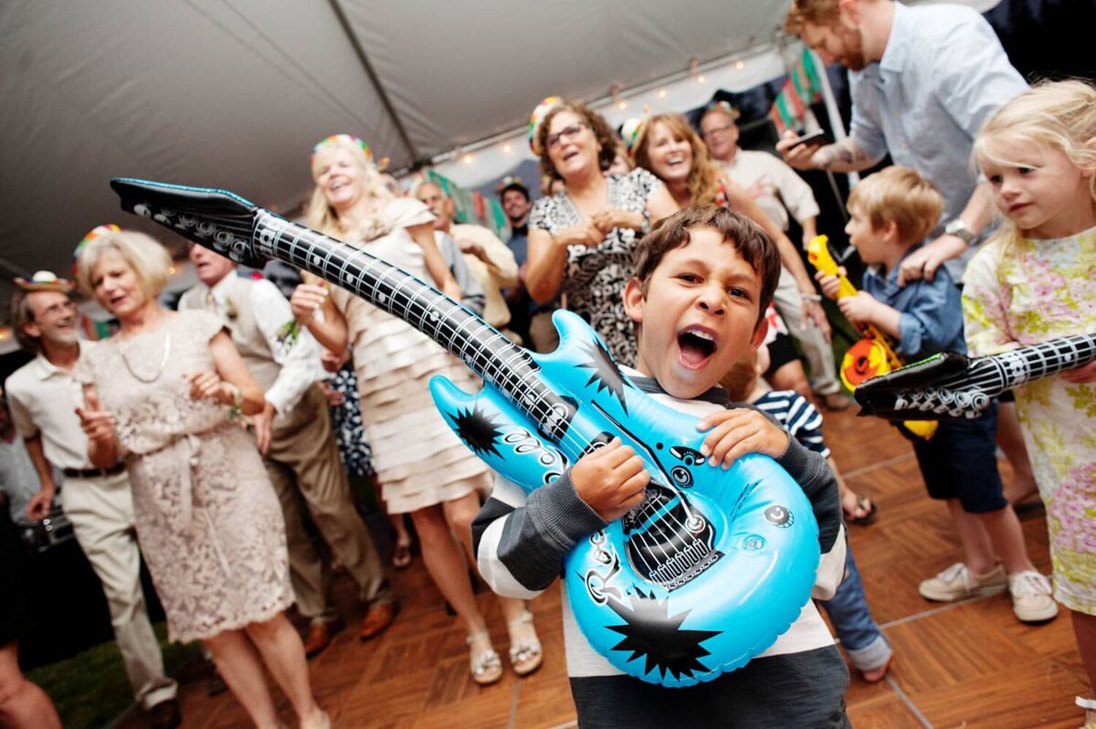 a little boy pretends to play a blow up guitar at a wedding reception with lots of people dancing in the background