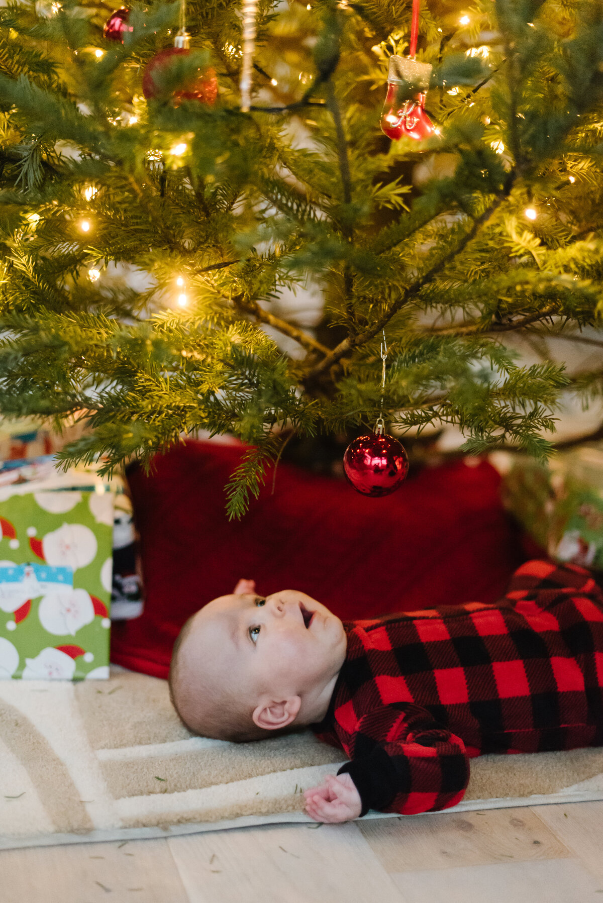Baby in red and black plaid onesie under a Christmas tree