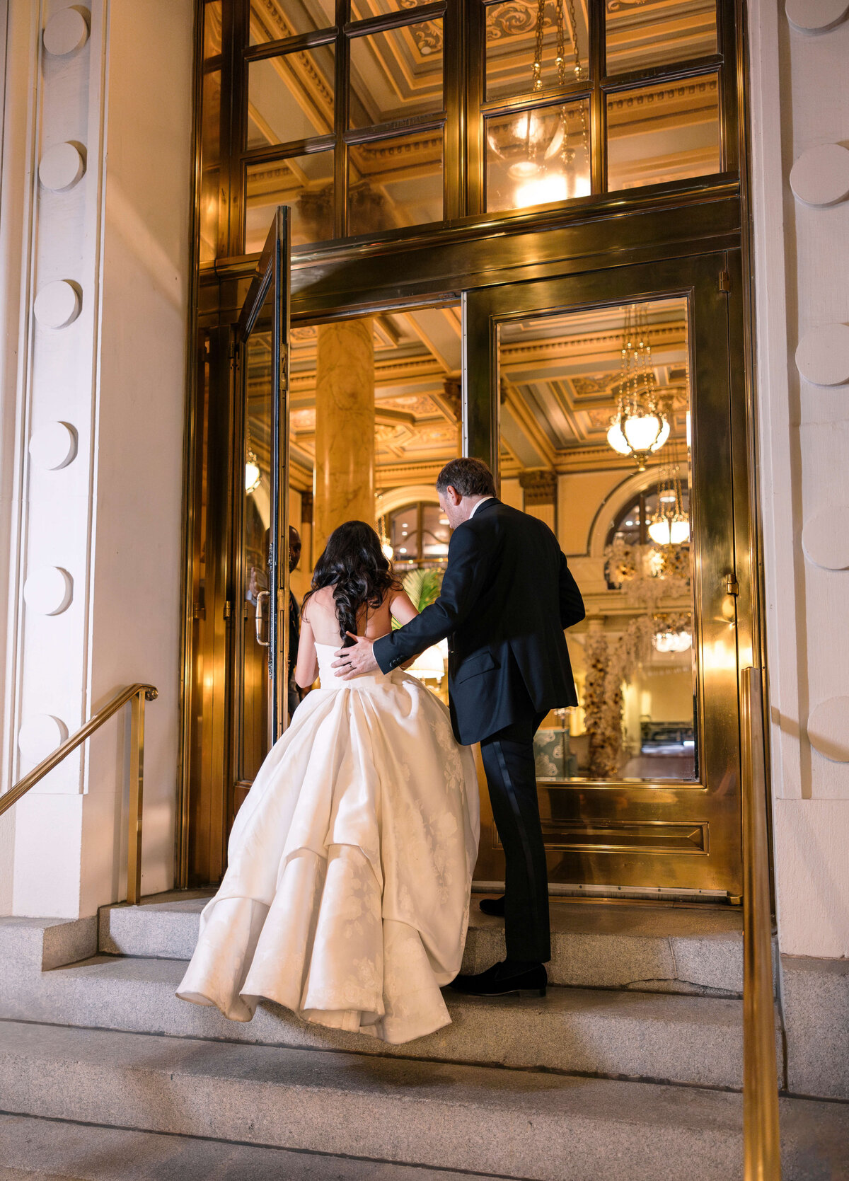 A couple elegantly dressed for a formal occasion ascends the steps to a building with golden doors. The woman wears a flowing white gown, and the man is in a black suit. The interior features ornate chandeliers and decorative elements.