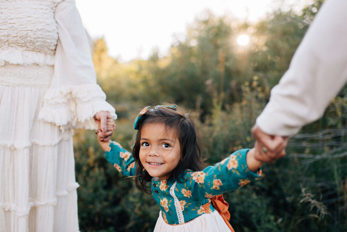 girl in blue dress at sunset in denver with arms outstretched holding mom and dad's hands
