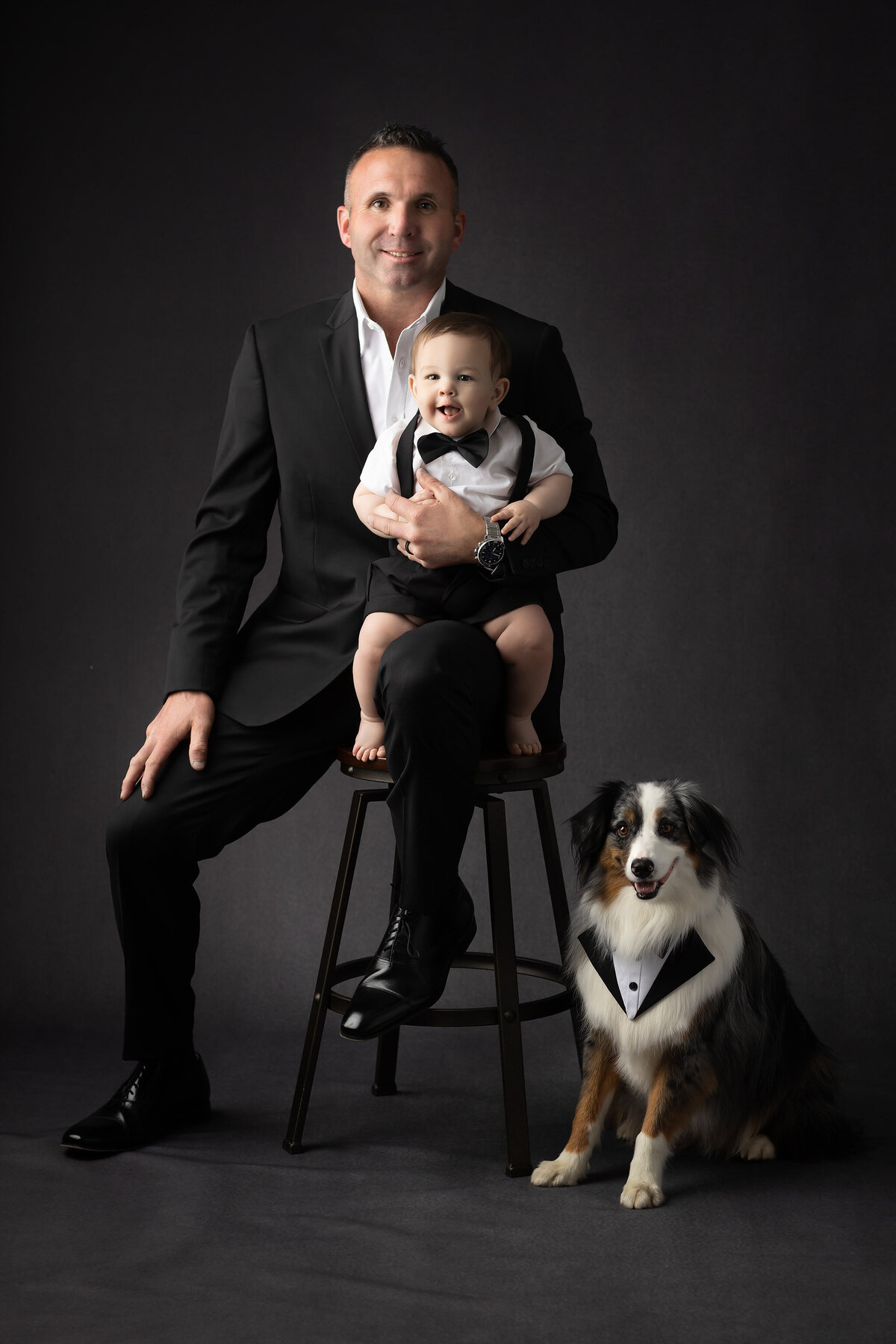 Milestone session featuring a father in a black suit holding his baby boy in suspenders and bow tie, with their tuxedo-clad dog beside them in a dark studio setting by a Dallas milestone photographer.