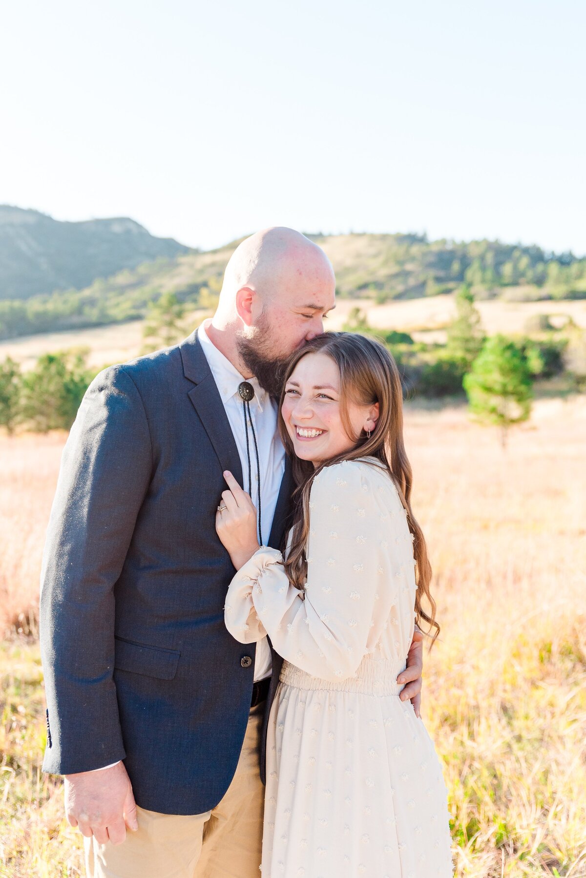 A woman in a tan long sleeved tan dress and long brown hair holds on to her spouses jacket lapel as he kisses her on the head with his eyes closed captured by denver family photographer