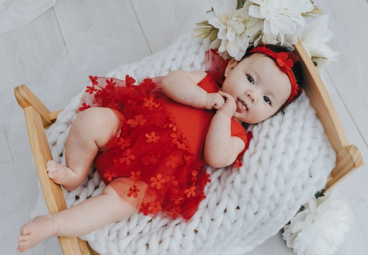 Baby girl in a bright red dress decorated with flower appliqués and matching headband, lying on a white chunky knit blanket with white flowers.