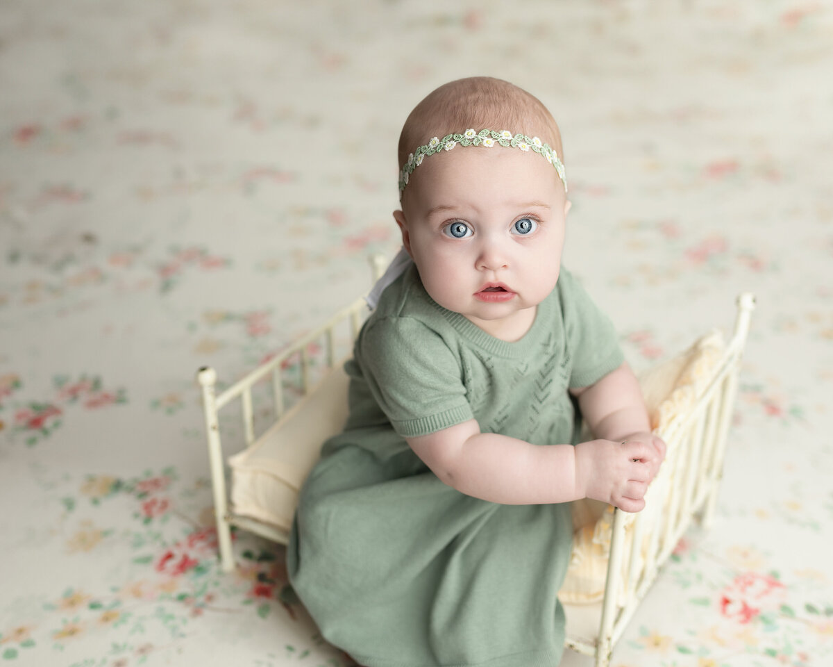 Vintage-inspired milestone of baby girl in sage dress sitting in a floral mini bed—styled softly with pastel tones.