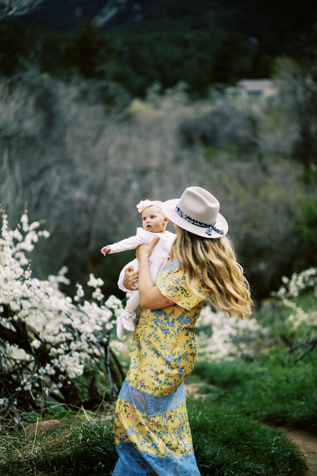 woman with long blonder hair twirls around on grass while holding her baby girl