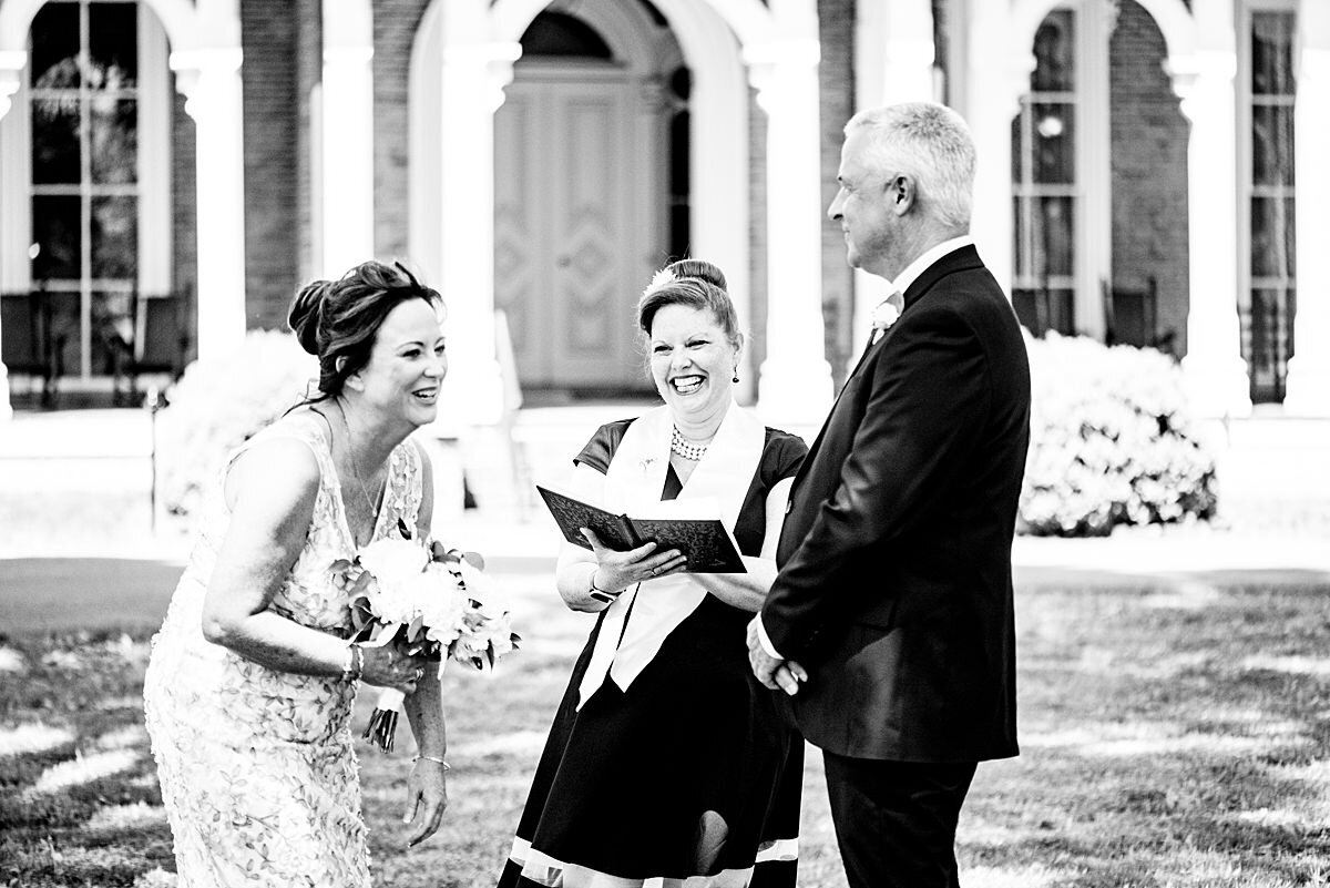 A black and white photo of the bride and groom laughing at something the officiant says as they stand on the lawn in front of  Oaklands Mansion. The bride is wearing a fitted lace dress and is holding a bouquet of white flowers. The officiant is wearing a black dress with a white officiant sash. She is holding a leather bound book. The groom is wearing a dark suit.
