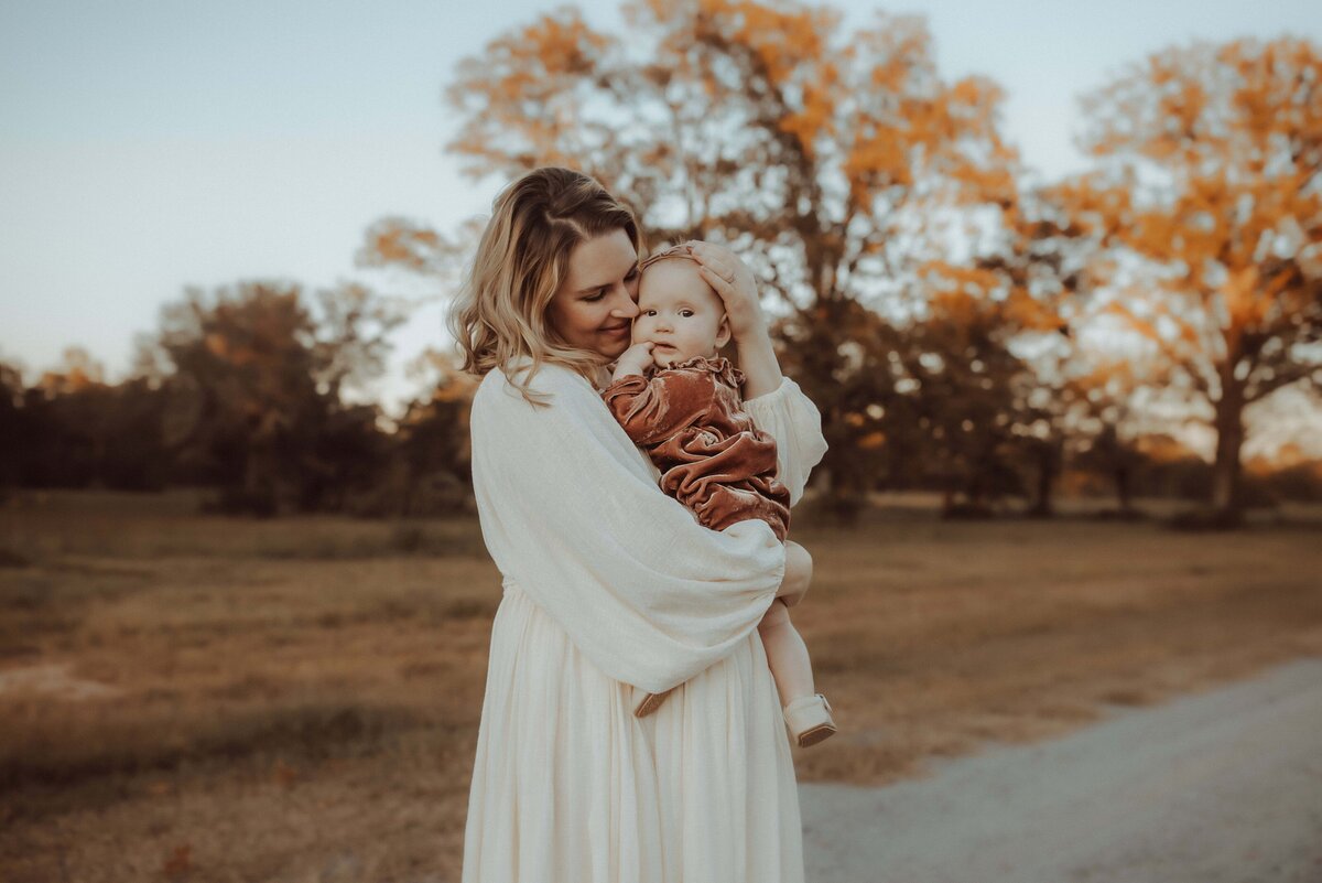 A woman in a white dress holds a baby wearing a rust-colored outfit. They stand on a dirt path with trees in the background under a clear sky.