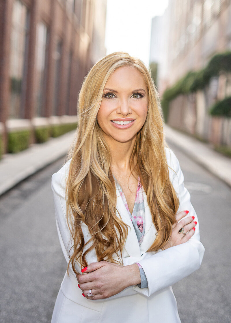 A young professional with blonde hair and a white blazer poses in the the street of the historic downtown Charleston cityscape.