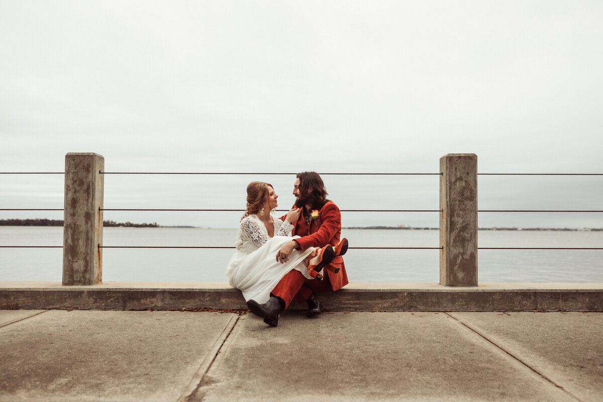 newlyweds sit side-by-side wearing retro vintage inspired wedding clothes - smiling at each other.  Portrait taken in the Cove in Panama City Florida