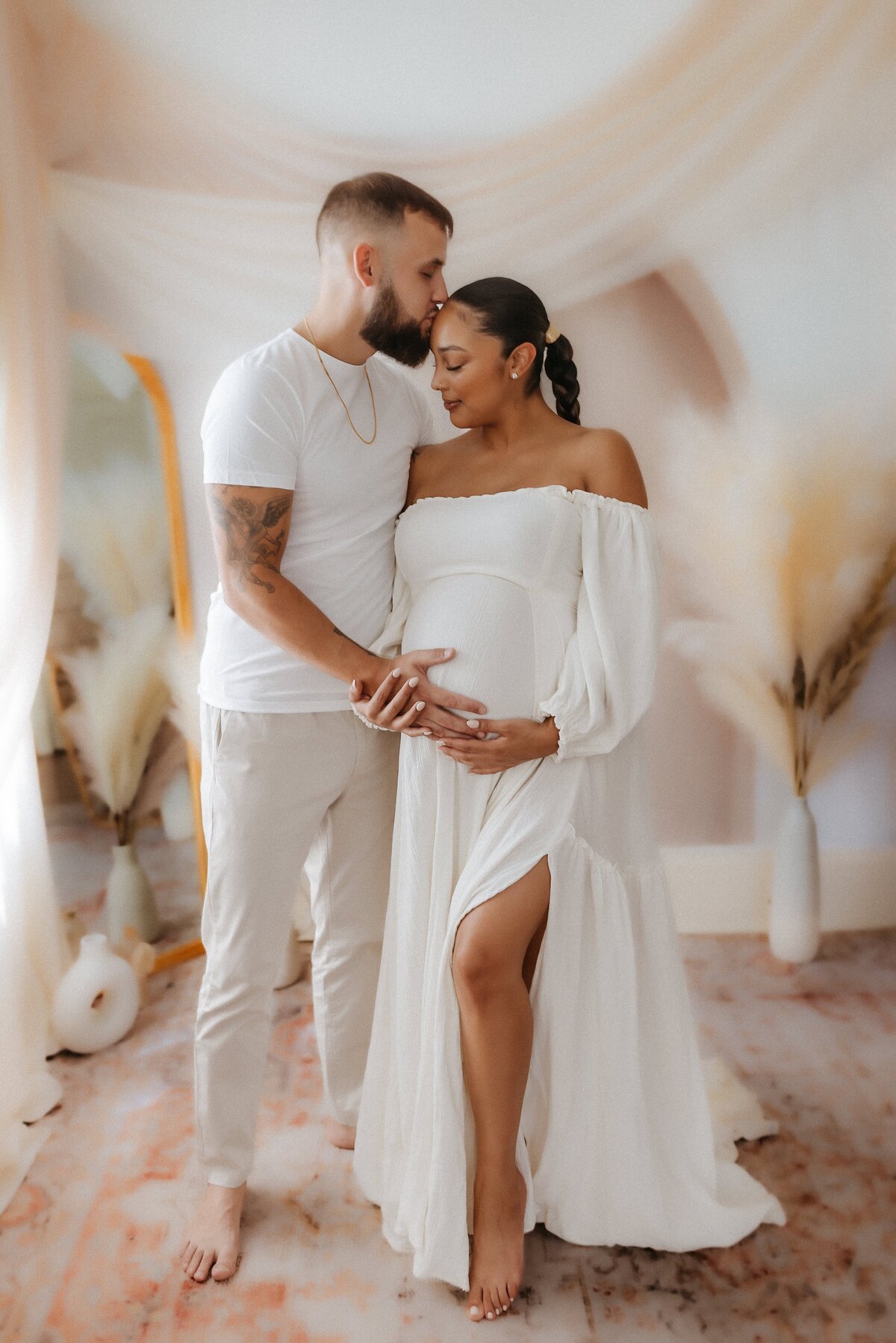 A man and a pregnant woman stand together in a softly lit room. The man is kissing the woman's forehead while both touch her belly. They are dressed in white.